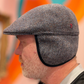 A man in profile wearing a gray herringbone tweed flat cap with ear flaps and black trim, set against a colorful pink, orange, and teal background.