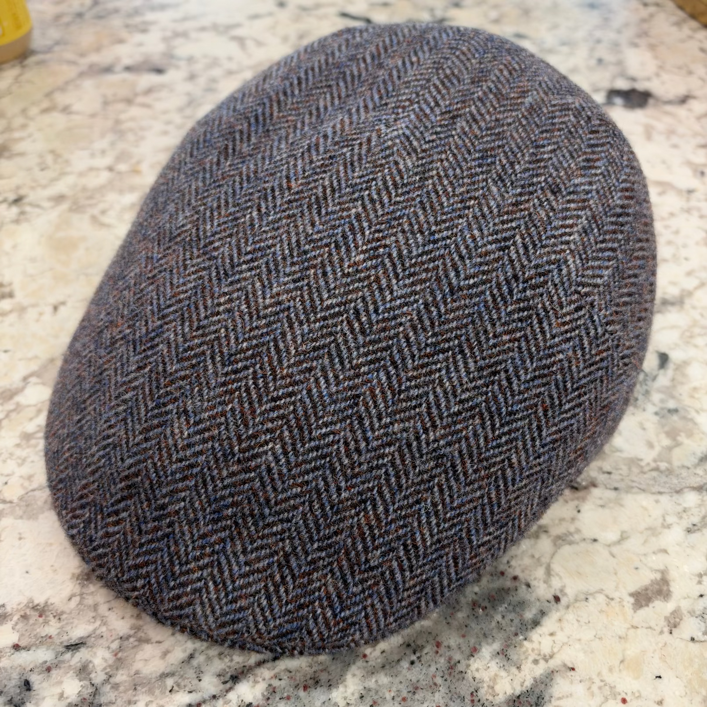 A classic flat cap in a brown and gray herringbone tweed pattern, resting on a speckled stone countertop.