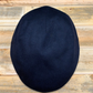 A dark navy blue wool flat cap, also known as a newsboy cap, shown from a top-down view on a rustic wooden plank background.