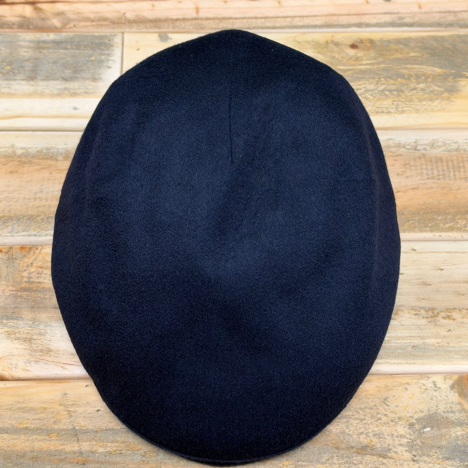 A dark navy blue wool flat cap, also known as a newsboy cap, shown from a top-down view on a rustic wooden plank background.