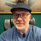 A close-up portrait of a man wearing a dark navy blue flat cap, black-rimmed eyeglasses, and a textured blue waffle-knit sweater, seated in a black leather office chair against a green wall.