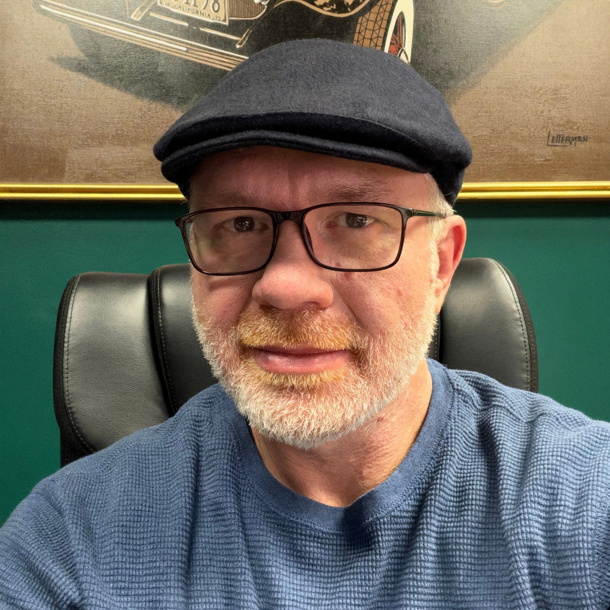A close-up portrait of a man wearing a dark navy blue flat cap, black-rimmed eyeglasses, and a textured blue waffle-knit sweater, seated in a black leather office chair against a green wall.