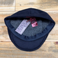 A dark navy blue knit scally cap with a gray fleece lining, displayed on a rustic wooden surface. The tags are attached.