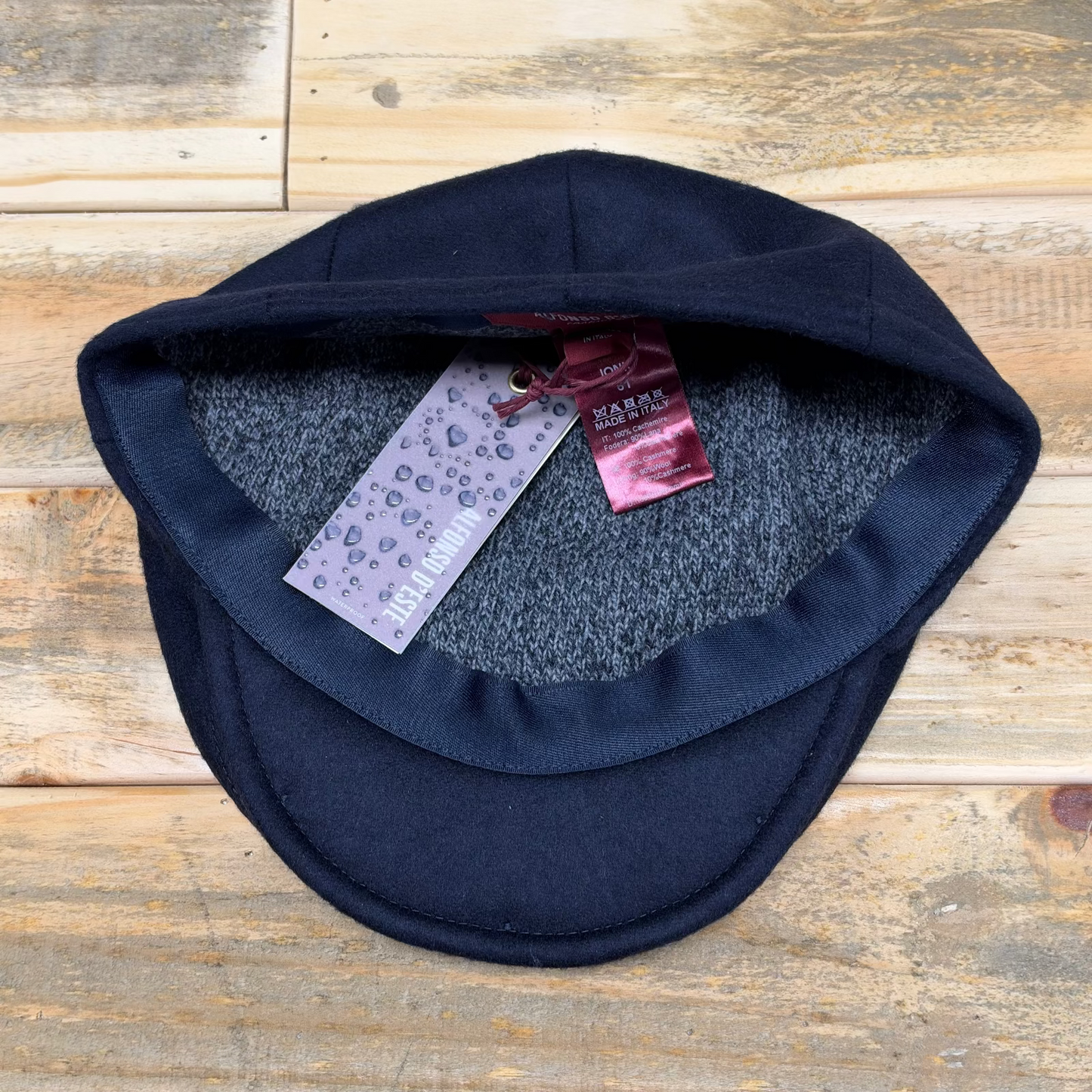 A dark navy blue knit scally cap with a gray fleece lining, displayed on a rustic wooden surface. The tags are attached.