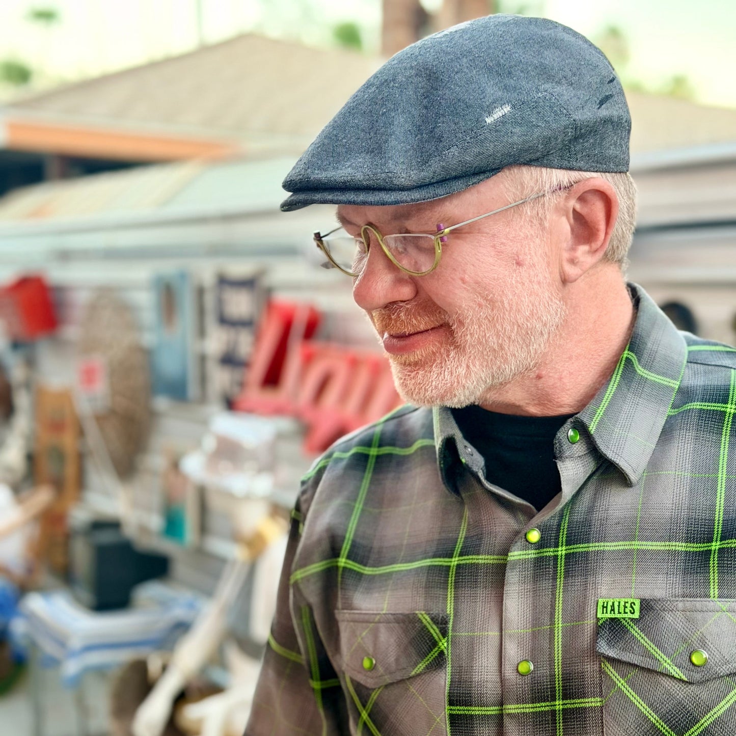 A man wearing a gray flat cap, wire-rimmed glasses, and a gray plaid short-sleeve button-up shirt with neon green lines and buttons. The shirt has a small green 'HALES' brand tag on the chest pocket.