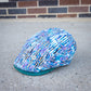A flat cap with an abstract multicolor print in blue, purple, white, and black, featuring a solid teal green visor, resting on a concrete surface with a brick wall in the background.
