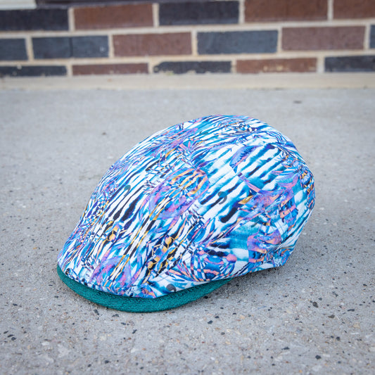 A flat cap with an abstract multicolor print in blue, purple, white, and black, featuring a solid teal green visor, resting on a concrete surface with a brick wall in the background.