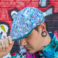 A man wearing a blue and white patterned flat cap with a dark green brim, posed in front of a graffiti-covered brick wall.