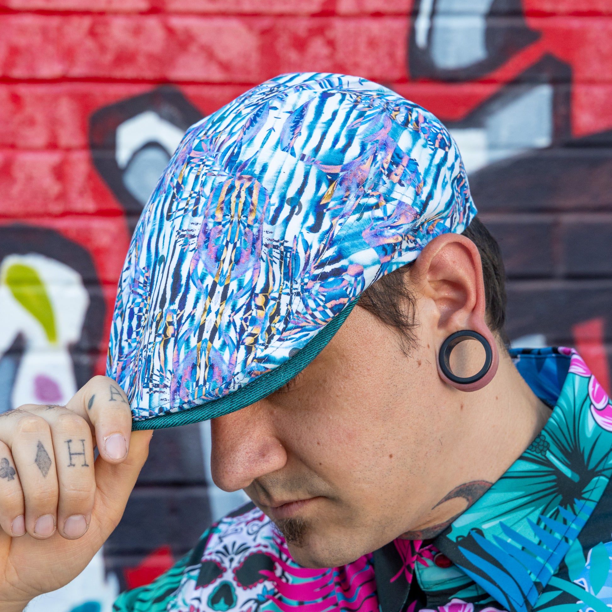 A man wearing a blue and white patterned flat cap with a dark green brim, posed in front of a graffiti-covered brick wall.