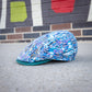 A flat cap with a vibrant blue, purple, and white abstract pattern, resting on a gray concrete surface with a brick wall and painted wall in the background.