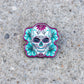 A sugar skull enamel pin with teal and magenta floral details, placed on a textured gray concrete surface.