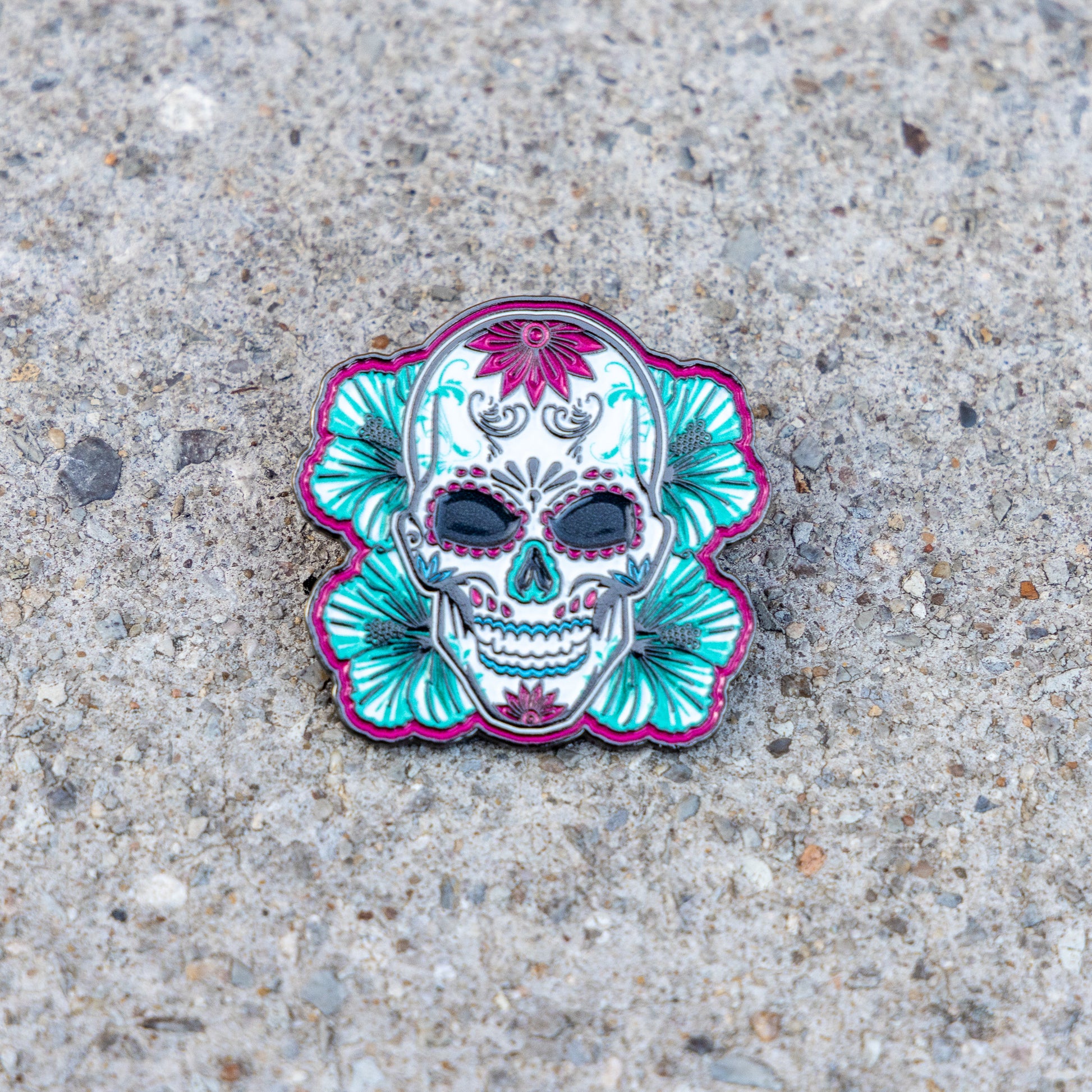 A sugar skull enamel pin with teal and magenta floral details, placed on a textured gray concrete surface.