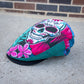 A flat cap with a vibrant sugar skull and floral print in pink, teal, white, and black, resting on a concrete surface with a brick wall in the background.