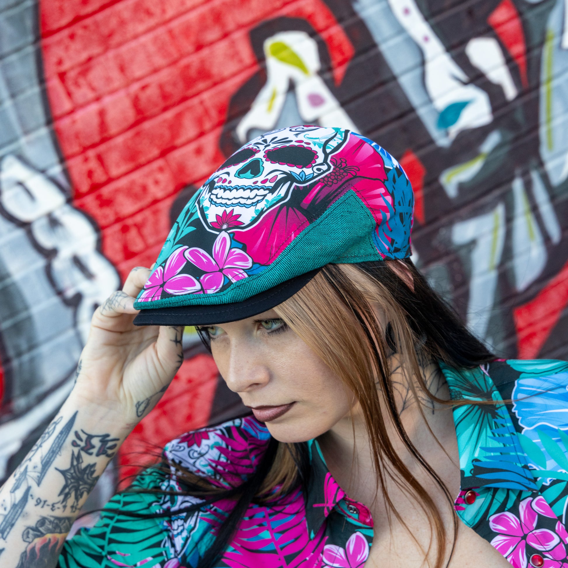 A woman wearing a colorful tropical and sugar skull print flat cap, posing in front of a vibrant graffiti wall.