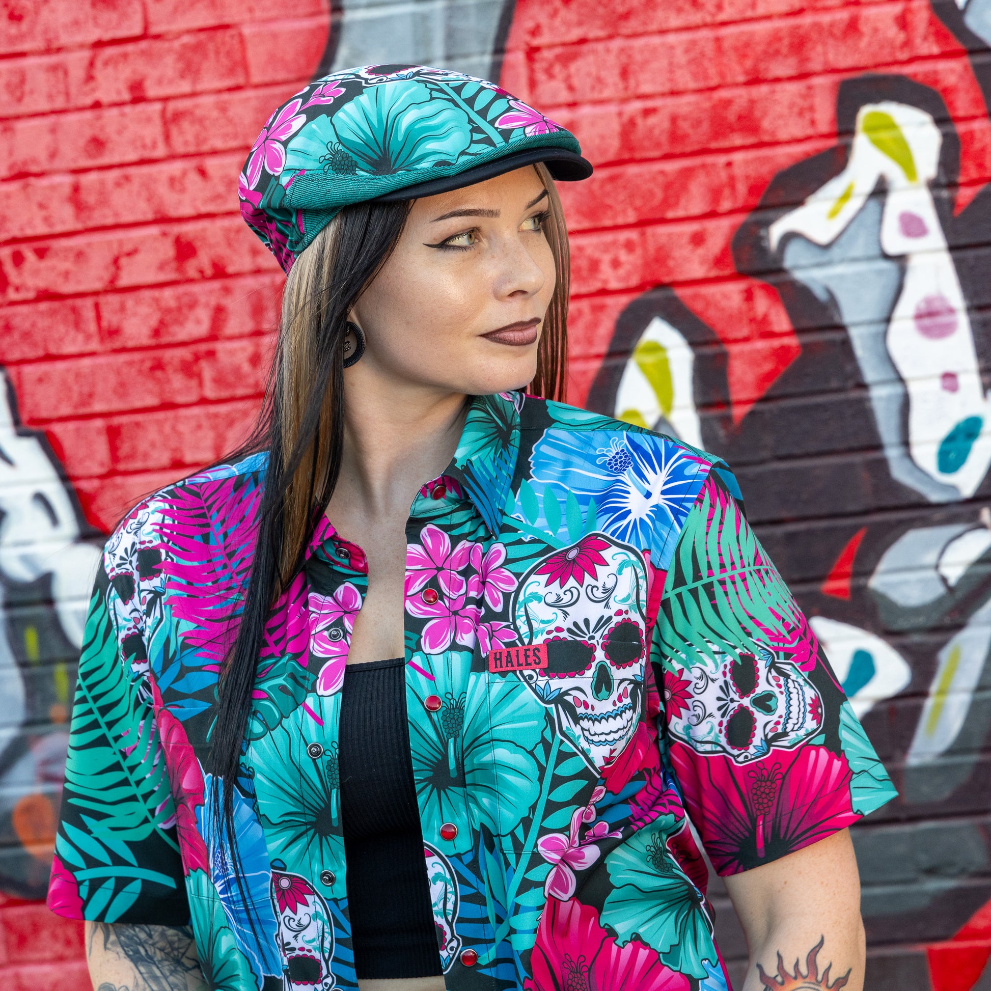 A woman wearing a vibrant tropical and sugar skull print short-sleeve button-up shirt and matching flat cap, posed in front of a graffiti-covered red brick wall.