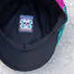 Close-up of the inside of a black cap showing a colorful embroidered patch with a skull with a teal mesh side panel visible on a concrete surface.