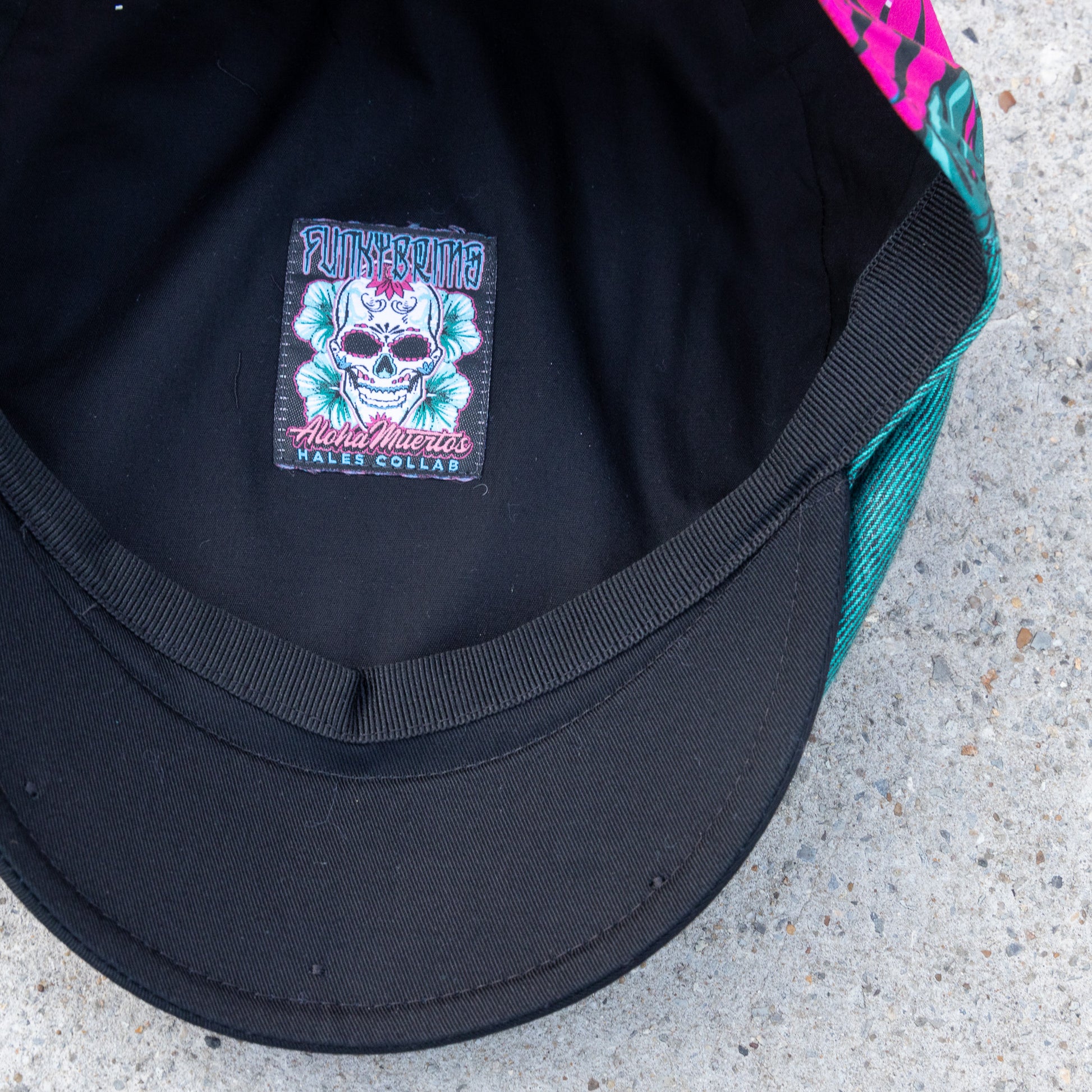 Close-up of the inside of a black cap showing a colorful embroidered patch with a skull with a teal mesh side panel visible on a concrete surface.
