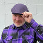 A man wearing a purple and black herringbone flat cap and a matching purple and black plaid button-down shirt, standing against a light gray painted brick wall.
