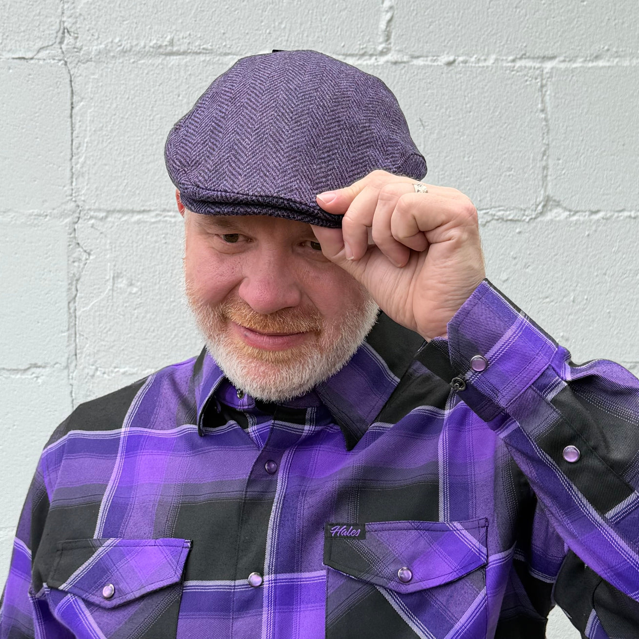 A man wearing a purple and black herringbone flat cap and a matching purple and black plaid button-down shirt, standing against a light gray painted brick wall.