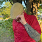 A man outdoors wearing a red button-up shirt and a tan cap, with his hand covering part of his face. The shirt has a small visible HUK logo on the chest pocket.
