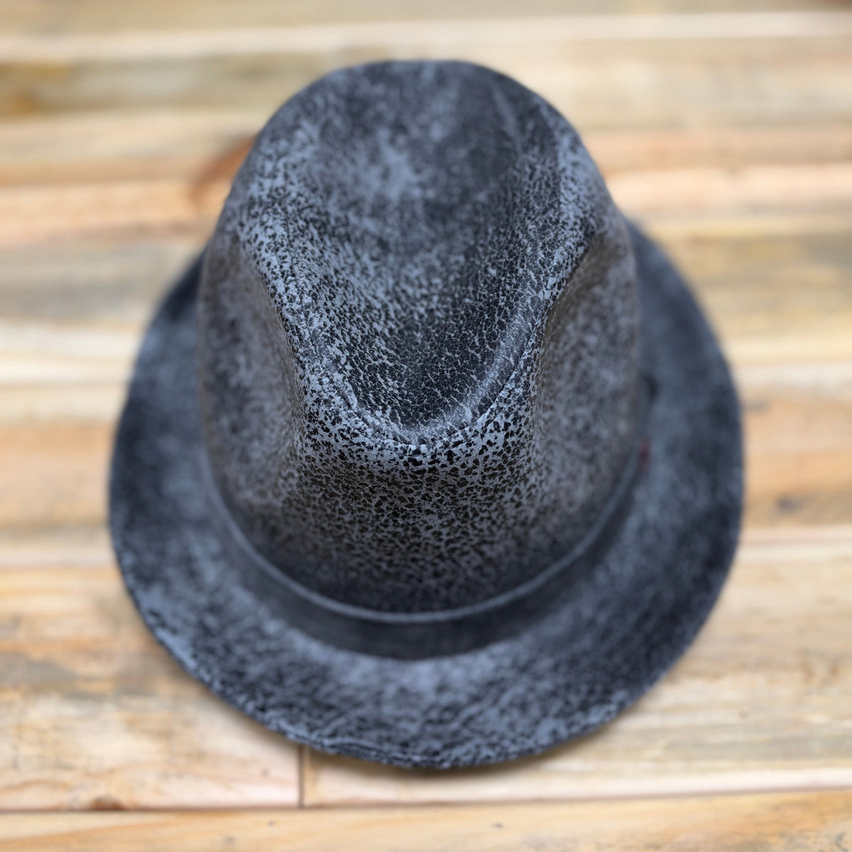 A dark gray textured fedora hat with a wide brim, photographed from a top-down perspective on a light brown wooden surface.