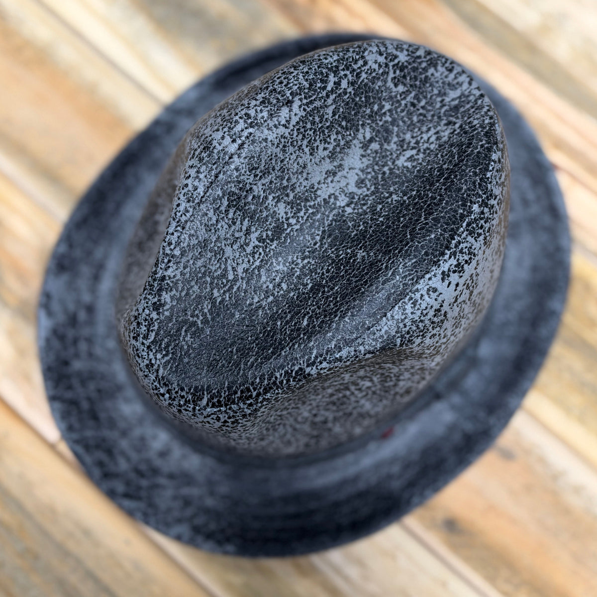 A black felt fedora hat with a textured, speckled finish, photographed from a top-down angle on a light-colored wooden plank surface.