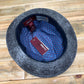 A top-down view of a dark gray felt hat with a blue and red patterned inner lining, resting on a light-colored wooden surface. The brand name 'FunkyBris' is visible on the tags.