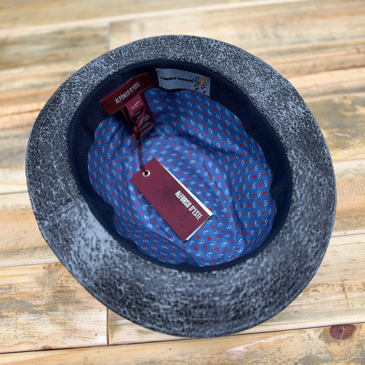 A top-down view of a dark gray felt hat with a blue and red patterned inner lining, resting on a light-colored wooden surface. The brand name 'FunkyBris' is visible on the tags.