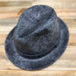 A black and gray speckled fedora hat with a black band, photographed from a top-down angle on a light wood background.