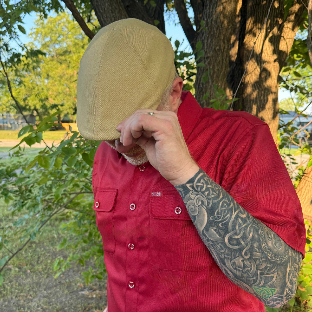 A man outdoors wearing a red button-up shirt and a tan cap, with his hand covering part of his face. The shirt has a small visible HUK logo on the chest pocket.