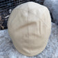 A beige flat cap made of canvas or twill fabric, photographed outdoors on a stone surface with a wire mesh fence in the background.