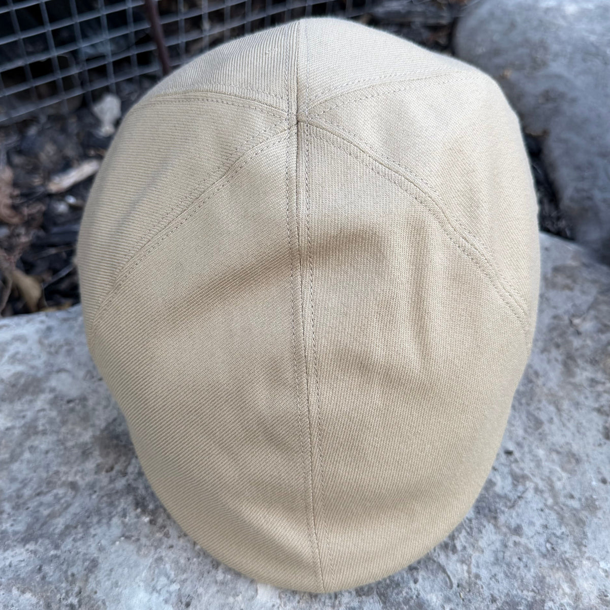 A beige flat cap made of canvas or twill fabric, photographed outdoors on a stone surface with a wire mesh fence in the background.