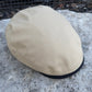 A beige canvas flat cap resting on a textured gray stone surface with a black fabric band visible along the edge.