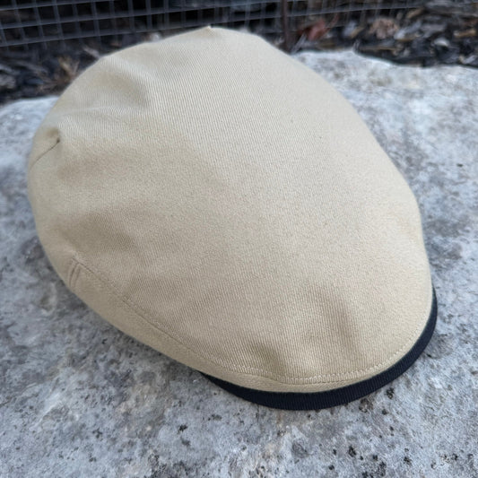 A beige canvas flat cap resting on a textured gray stone surface with a black fabric band visible along the edge.