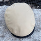 A beige flat cap with a black ribbed trim band, resting on a textured gray stone surface with a wire mesh fence in the background.