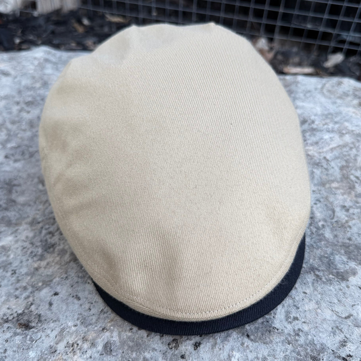 A beige flat cap with a black ribbed trim band, resting on a textured gray stone surface with a wire mesh fence in the background.