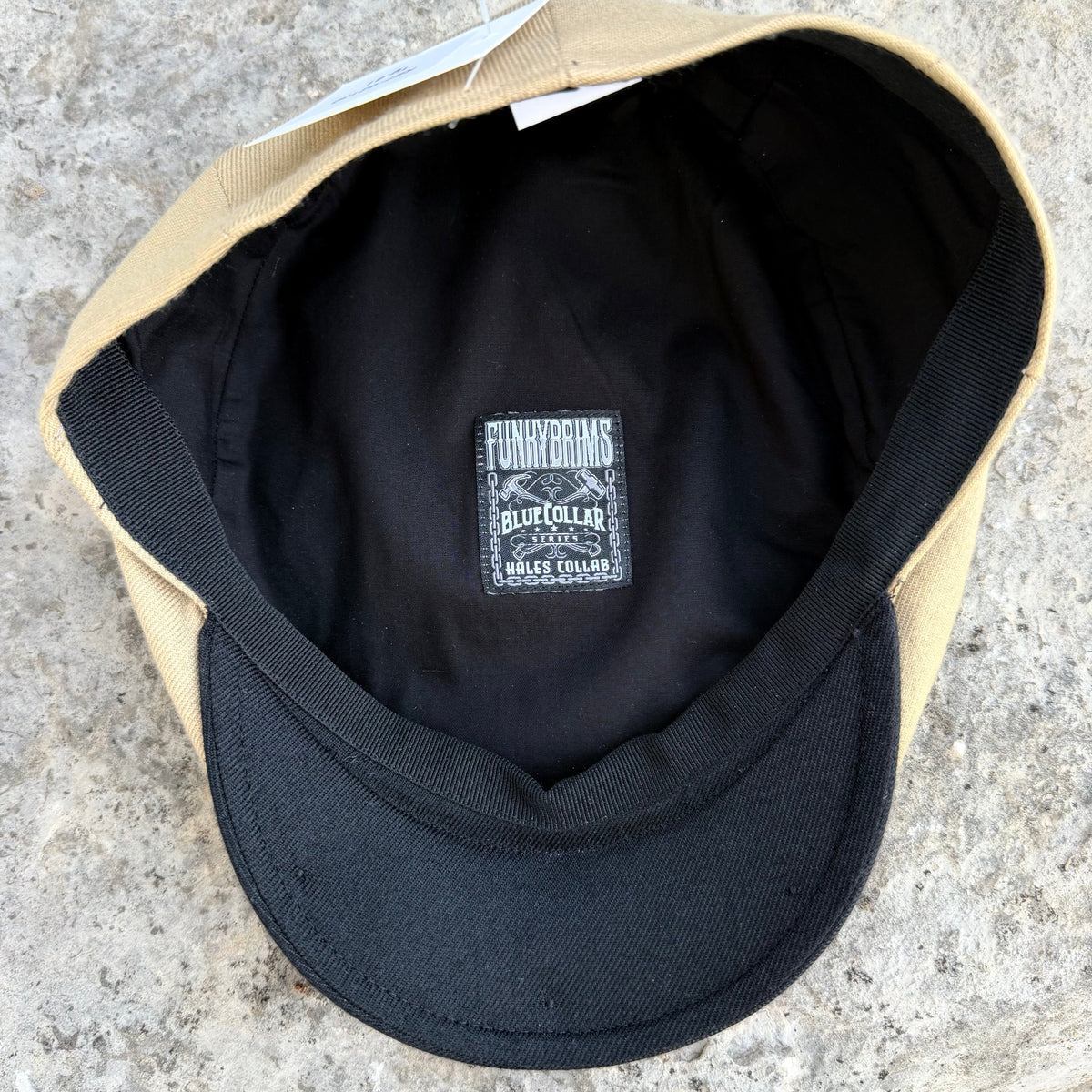 A close-up top-down view of a baseball cap with a tan crown and black visor, showing the black interior lining and a central sewn label that reads 'FLYING BRIMS BLUE COLLAR'.