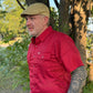 A man wearing a red short-sleeve button-up shirt and a tan flat cap, standing outdoors in front of trees.