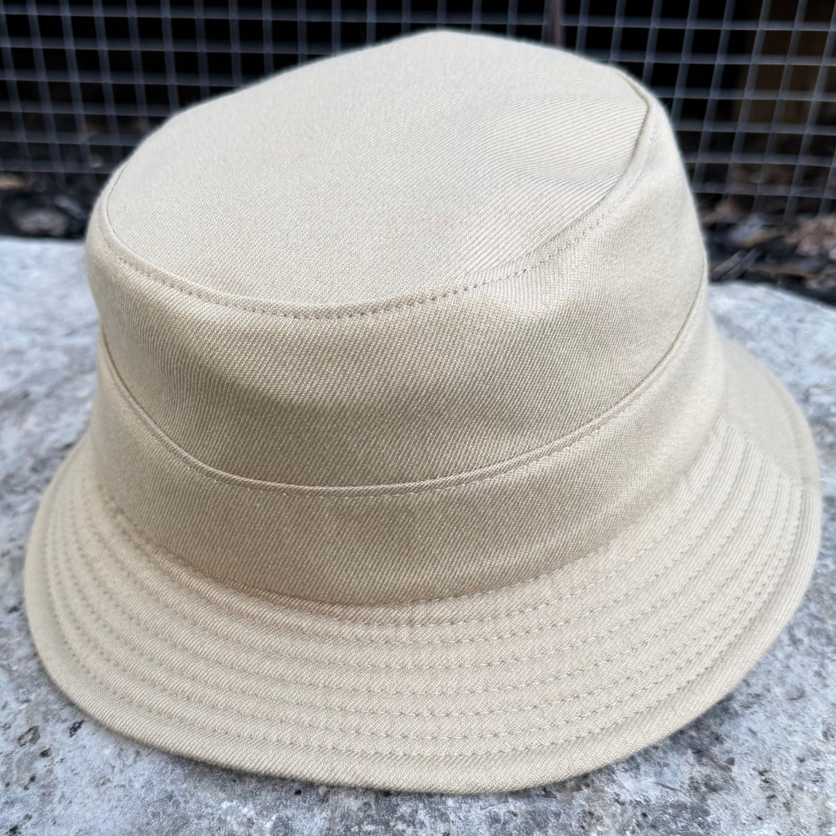 A light beige canvas bucket hat resting on a textured gray stone surface with a wire mesh fence in the background.