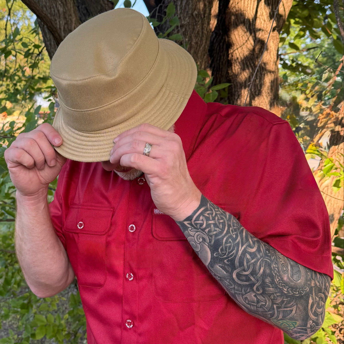 A man wearing a tan bucket hat and a red short-sleeve button-up shirt, standing outdoors in front of trees and foliage.