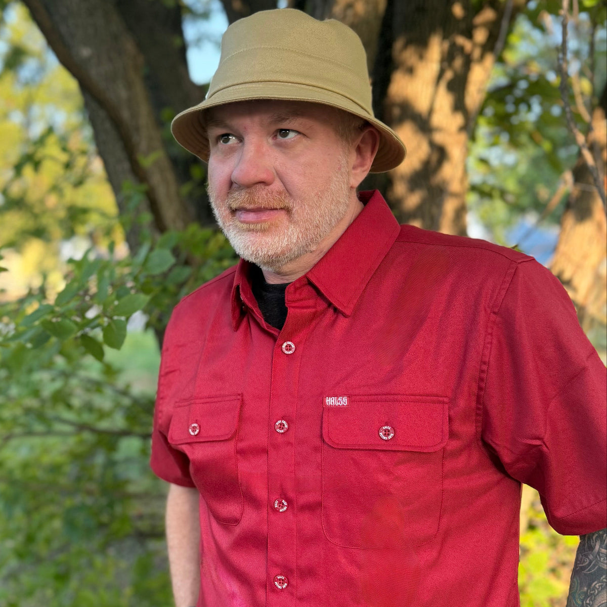 A man wearing a red short-sleeve button-up shirt and a tan bucket hat, standing outdoors in front of trees.