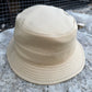 A beige canvas bucket hat with a stitched brim, resting on a textured gray stone surface with a dark metal grid fence in the background.