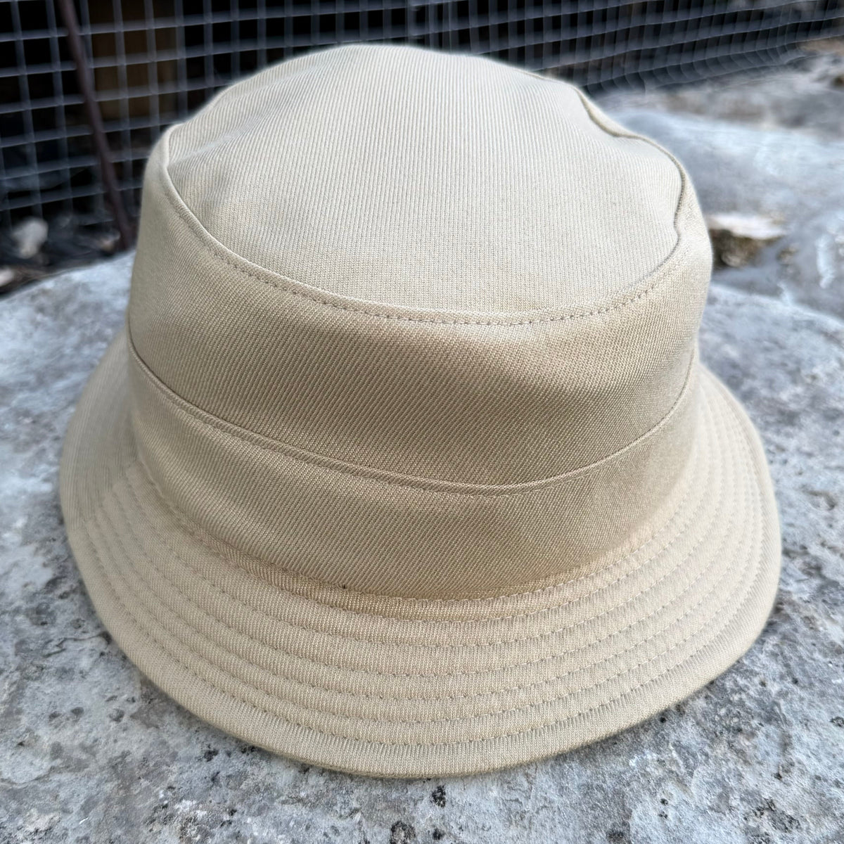 A beige canvas bucket hat with a stitched brim, resting on a textured gray stone surface with a dark metal grid fence in the background.