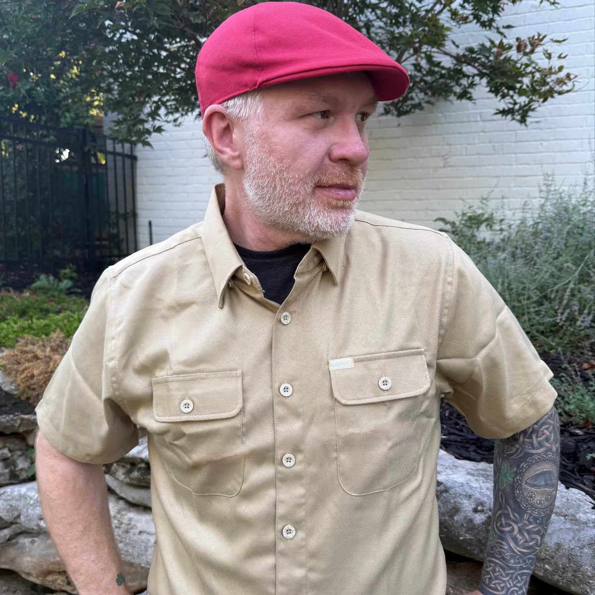 A man wearing a beige short-sleeve button-up shirt with two chest pockets and a red flat cap, standing outdoors in front of a white brick wall and garden plants.