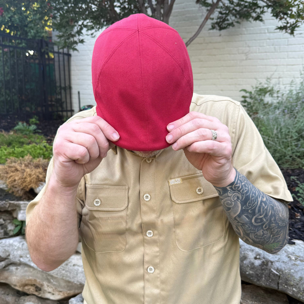 A man wearing a beige short-sleeve button-up shirt and a bright red baseball cap, holding the cap in front of his face with both hands in an outdoor backyard setting.