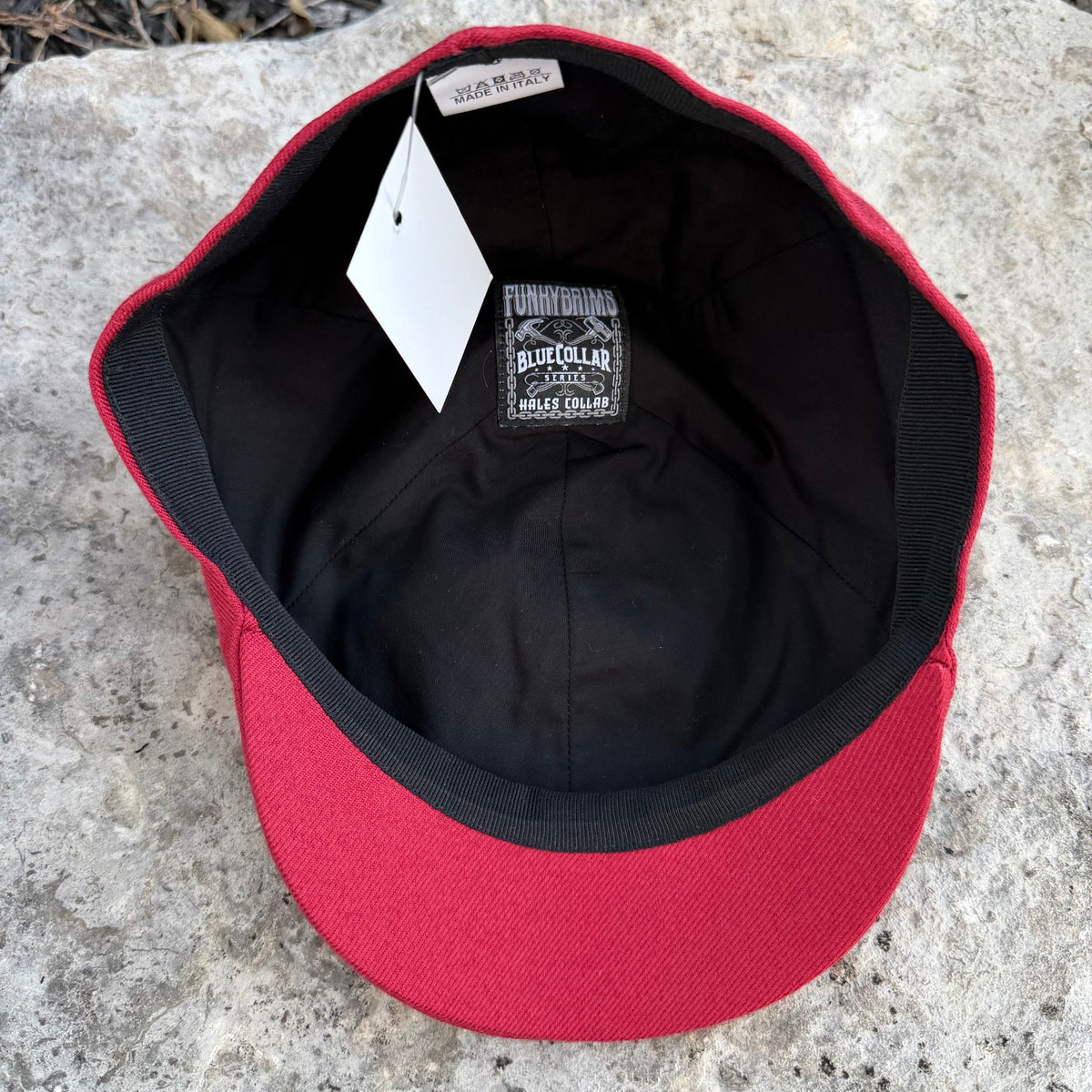 A red and black baseball cap shown from the inside, resting on a gray stone surface. The cap features a red crown and brim, a black interior, a black ribbed trim, a white hangtag, and a black interior label reading 'Blue Collar Caps'.
