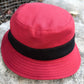 A red bucket hat with a black ribbon band, photographed outdoors on a stone surface.