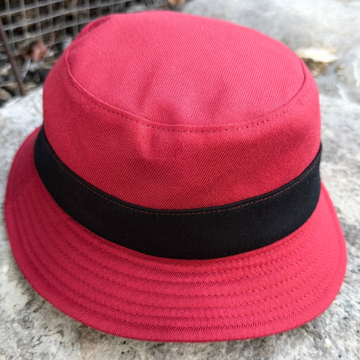 A red bucket hat with a black ribbon band, photographed outdoors on a stone surface.