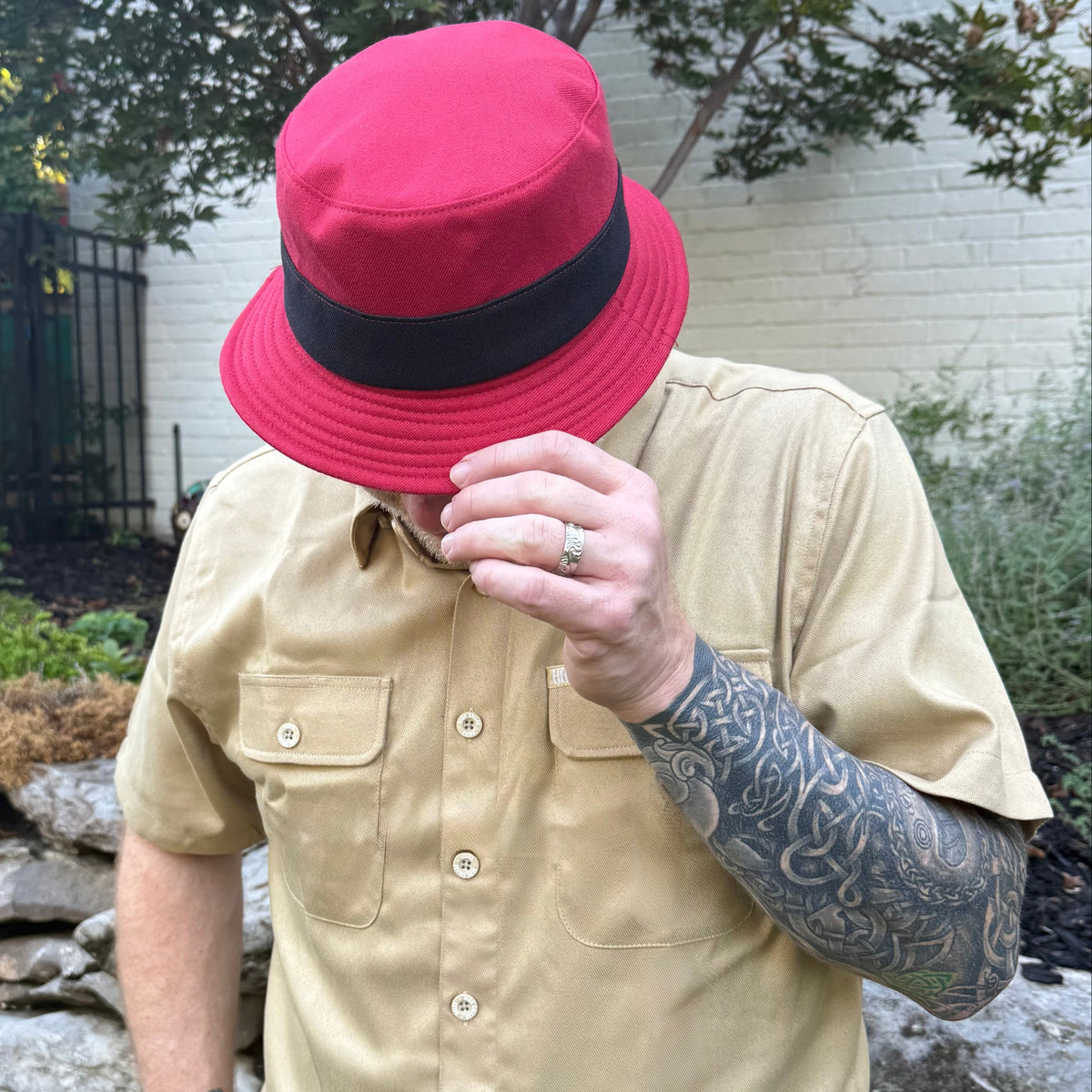 A man wearing a red fedora-style hat with a black band, a beige short-sleeve button-up shirt, and a tattooed arm, standing outdoors in front of a white brick wall.