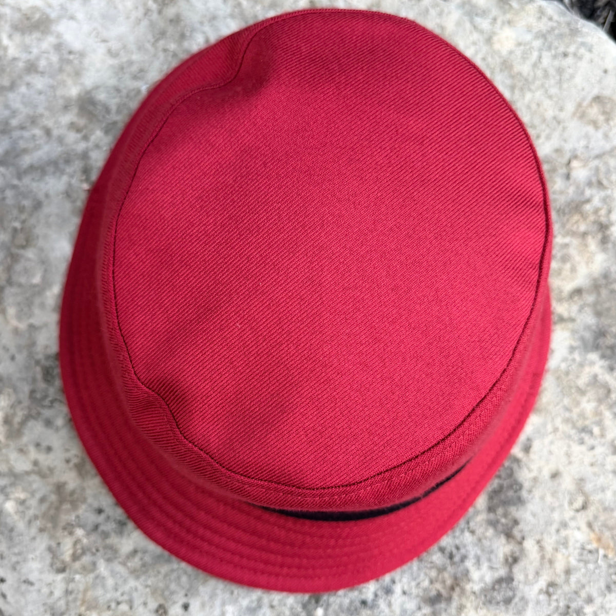A red bucket hat with a black band, photographed from a top-down angle on a textured gray stone surface.